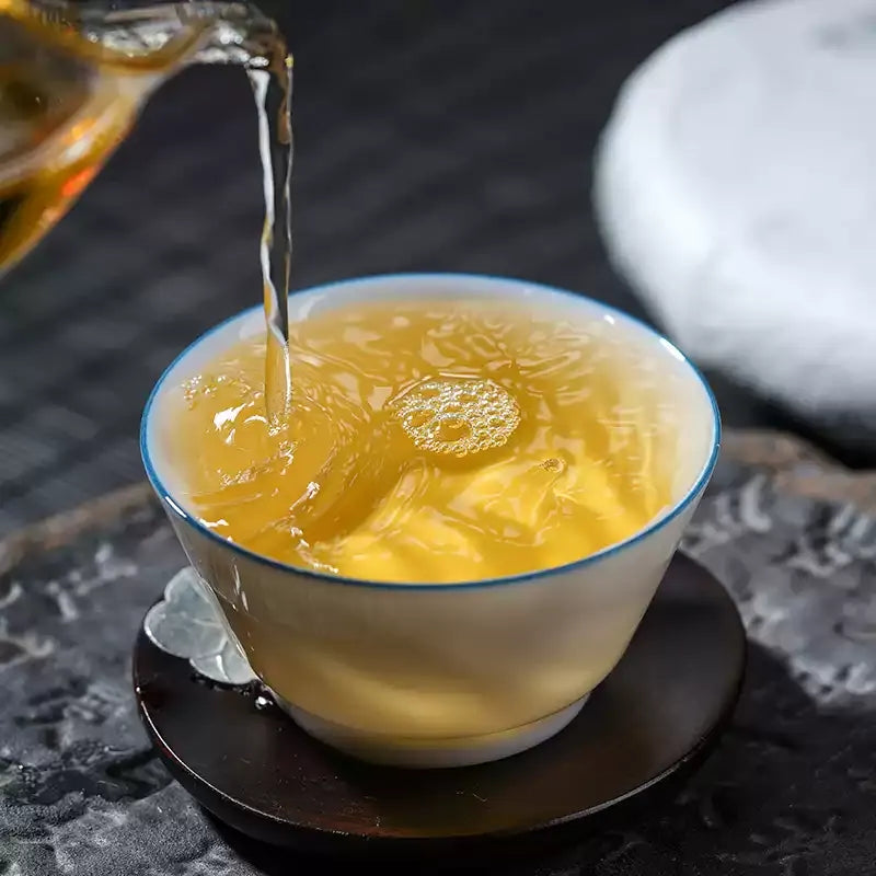 Golden-amber Yibang raw pu-erh tea liquor being poured into a white porcelain cup — 2025 Yunnan sheng puerh from the Six Famous Tea Mountains, smooth honeyed infusion